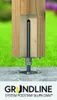 A close-up of a wooden post base with a metal threaded support system, set against a blurred green lawn and a concrete surface. The brand name and product description are displayed at the bottom. Produktbild-Vorschau
