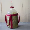A large clear plastic carboy filled with small green objects, possibly hops or fruit, with a red lid and an airlock. It has two pink straps for carrying. The carboy sits on a gray surface against a light wood-paneled background. Produktbild-Vorschau