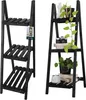 Two black wooden ladder shelves are shown side-by-side against a white background. The left shelf is empty except for a product tag. The right shelf displays several potted plants, books, and a vintage camera. Produktbild-Vorschau