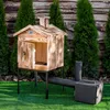 A wooden smoker oven with a metal chimney and a connected firebox, set up on a green lawn in front of a wooden fence and evergreen trees. Náhled obrázku produktu