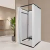 A freestanding changing room with white curtains and a black frame, set up in a retail store environment. Shelving and a plant are visible in the background. Produktbild-Vorschau