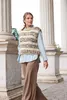 A woman walks past large stone columns, wearing a knitted vest over a light blue shirt and brown pants. Produktbild-Vorschau