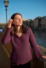 A woman wearing a knitted sweater stands on a bridge with a river and buildings in the background. Produktbild-Vorschau