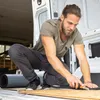 A man is crouching inside a van, cutting a piece of insulation material with a utility knife. Produktbild-Vorschau