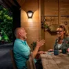 A couple is sitting at a wooden table outdoors, possibly on a patio or in a garden, engaged in conversation. A wall-mounted outdoor light fixture is visible on the wooden wall behind them. Anteprima dell’immagine prodotto