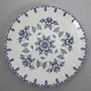 A white ceramic plate with a blue floral pattern is shown from a top-down perspective on a light gray textured surface. Produktbild-Vorschau
