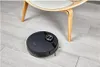 A black robot vacuum cleaner is on a light gray wood-look floor under a chair. Podgląd zdjęcia produktu