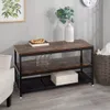 A three-tier storage shelf with a rustic wood top and black metal frame, decorated with a book, beads, and a vase with greenery. The shelf is placed in a room with a chair, a basket, and wall art visible. Náhled obrázku produktu