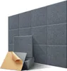 A wall of dark gray acoustic panels with a stack of panels and one panel with its adhesive backing exposed in the foreground. Produktbild-Vorschau