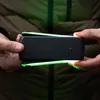 A person is holding a black electronic device with green lights emanating from the top and bottom. Voir les images du produit