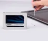 A Crucial MX500 SSD is shown on a white surface, with a hand using a screwdriver to work on a laptop in the background. Podgląd zdjęcia produktu
