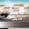 A pair of white Philips earbuds and their charging case are placed on a wooden table next to a cup of coffee, with a blurred background of colorful buildings and a blue sky. Podgląd zdjęcia produktu