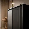 A close-up view of a black cabinet with vertical slats and a smooth door, decorated with a vase of dried grass and books. Produktbild-Vorschau