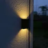 A black solar outdoor wall light is mounted on a textured wall, casting warm yellow light upwards and downwards. The background shows a dark, out-of-focus garden scene at dusk. Náhľad obrázku produktu