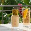 A perfume bottle and a drink with an orange slice on a table outdoors, with greenery in the background. Voir les images du produit