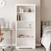 A white bookshelf filled with books and decorative items, placed in a room with a desk and a bed. Produktbild-Vorschau
