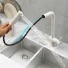 A person is using a white kitchen faucet with a pull-out spray head over a double stainless steel sink. Náhled obrázku produktu