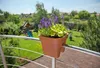 A planter box with purple flowers and green leaves is attached to a balcony railing, overlooking a backyard with a playground. Produktbild-Vorschau