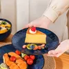 A person is serving a slice of cheesecake on a decorative blue plate, with other food items visible. Náhled obrázku produktu