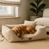 A dog is sleeping peacefully in a beige dog bed next to a window and a plant. Produktbild-Vorschau