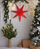 A red star-shaped Christmas decoration hangs on a white brick wall, with a small Christmas tree in a pot to the left and a larger decorated Christmas tree to the right. There are also gift boxes on the floor. Podgląd zdjęcia produktu