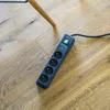 A power strip with four outlets sits on a wooden floor next to a chair leg. Produktbild-Vorschau