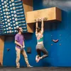 Eine Person hängt an einem Trainingboard namens 'VerticalBoard One' von YY Vertical; während eine andere Person zuschaut. Die Wand ist mit verschiedenen Klettergriffen ausgestattet. Produktbild-Vorschau