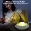 A mother is breastfeeding her baby at night, with a sound machine on a table. Produktbild-Vorschau