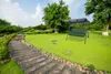 A garden swing with a green cushion is set up on a lush green lawn next to a stone pathway. Produktbild-Vorschau