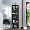 A tall, narrow black bookshelf filled with decorative items and books, next to a grey curtain and a window with greenery outside. A dark grey ottoman with a furry cushion is in the foreground. Náhled obrázku produktu