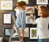 A mother and daughter are hanging children's artwork in frames on a dark wall. Produktbild-Vorschau
