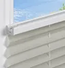 A close-up view of a light gray pleated blind installed on a window frame, with a blue sky and clouds visible through the window. Produktbild-Vorschau