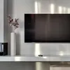 A modern living room setup with a TV mounted on a white wall, a white vase with decorative branches, and a remote control on a white shelf. Produktbild-Vorschau