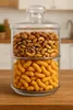 A two-tiered glass jar filled with nuts and cheese puffs, placed on a wooden table. Produktbild-Vorschau