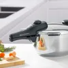 A close-up shot of a pressure cooker with carrots and herbs in the foreground. Náhled obrázku produktu