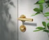 A close-up shot of a gold door handle and lock on a white door, with blurred green leaves in the background. Produktbild-Vorschau