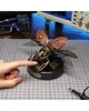 A hand is touching a mechanical rhinoceros beetle model on a grid mat, with electronic equipment in the background. Náhľad obrázku produktu