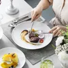 A person is eating a meal with steak and vegetables from a white plate, with a slice of cheesecake on another plate in the foreground. Produktbild-Vorschau