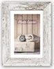 A vintage-style alarm clock and a folded blanket rest on a cream-colored suitcase, set against a backdrop of vertical wooden planks. The scene is framed by a distressed white wooden picture frame. Podgląd zdjęcia produktu
