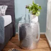 A silver hammered metal side table with a plant, a glass of water, and a carafe of water on top, next to a bed. Náhled obrázku produktu