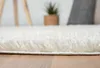 A close-up view of a fluffy white rug on a wooden floor, with a blurred background of a plant and pillows. Náhľad obrázku produktu