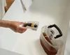 A person is holding a small, clear plastic container filled with debris, possibly hair and dirt, over a larger, clear plastic water tank with a black handle. The items are placed on a white surface, likely a sink or countertop. Podgląd zdjęcia produktu