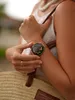 A woman's arm wearing a rose gold smartwatch with a floral design on the face, holding a straw bag. Anteprima dell’immagine prodotto