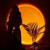 A person stands in front of a sunset projection lamp, casting a shadow on the wall. A plant is in the foreground. Produktbild-Vorschau