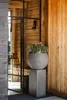 A planter on a pedestal in a rustic entryway with wooden walls and a large glass door. Produktbild-Vorschau