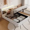 A white bed with its storage compartment open, revealing stored items. The bed has a tufted headboard with LED lighting. Náhled obrázku produktu