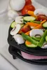 A close-up shot of a black cast iron grill insert filled with various chopped vegetables, including mushrooms, zucchini, carrots, tomatoes, and green peppers, placed on top of a pink patty. Produktbild-Vorschau