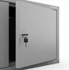 A close-up view of a gray metal cabinet with one door slightly ajar, revealing a dark interior. A key is attached to the lock on the open door. Produktbild-Vorschau
