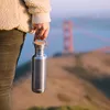A person holding a stainless steel water bottle with a bamboo lid, with the Golden Gate Bridge in the background. Podgląd zdjęcia produktu