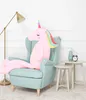 A large pink unicorn plush toy sits in a light blue armchair next to a small side table with a vase and an alarm clock. Produktbild-Vorschau